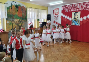 Dziewczynki w białych sukienkach z czerwonymi paskami oraz chłopcy w białych koszulach, czerwono-czarnych kamizelkach oraz czarnych spodniach tańczą Poloneza.